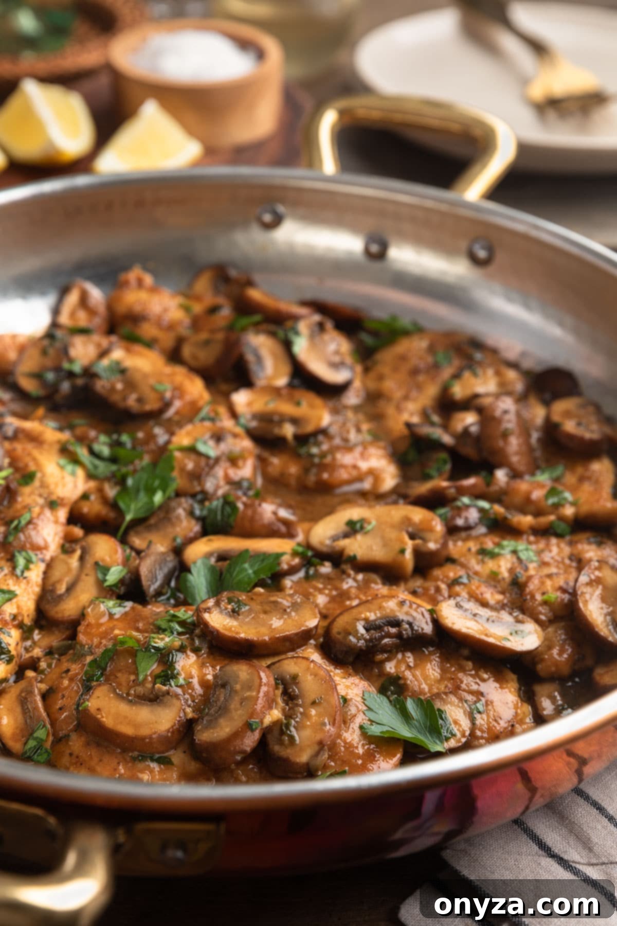 A close-up side view of a serving of Chicken Marsala artfully arranged in a rustic copper frying pan, showcasing the rich sauce and tender chicken.