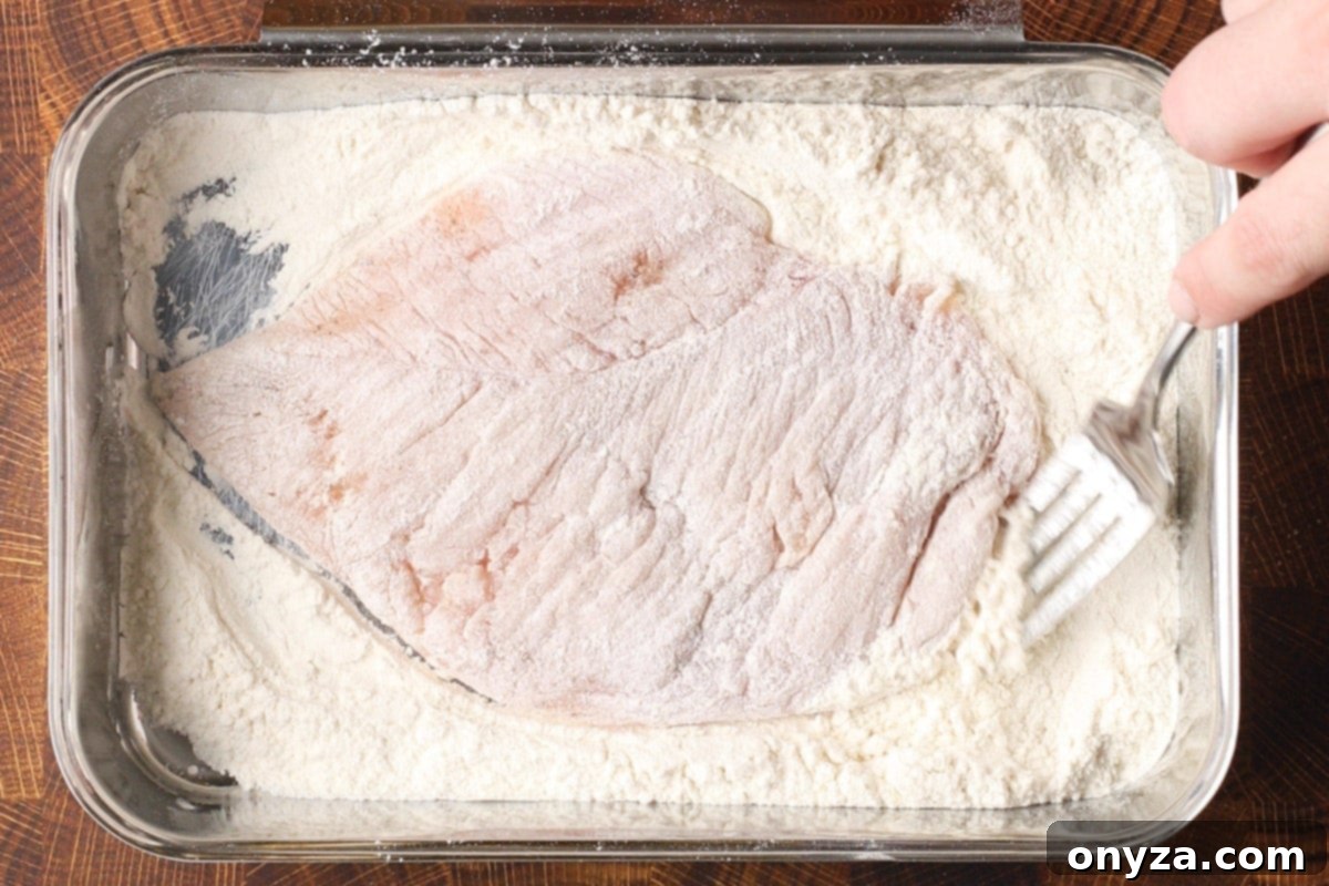 A chicken cutlet being carefully dredged in a shallow dish of all-purpose flour, ensuring an even coating before cooking.