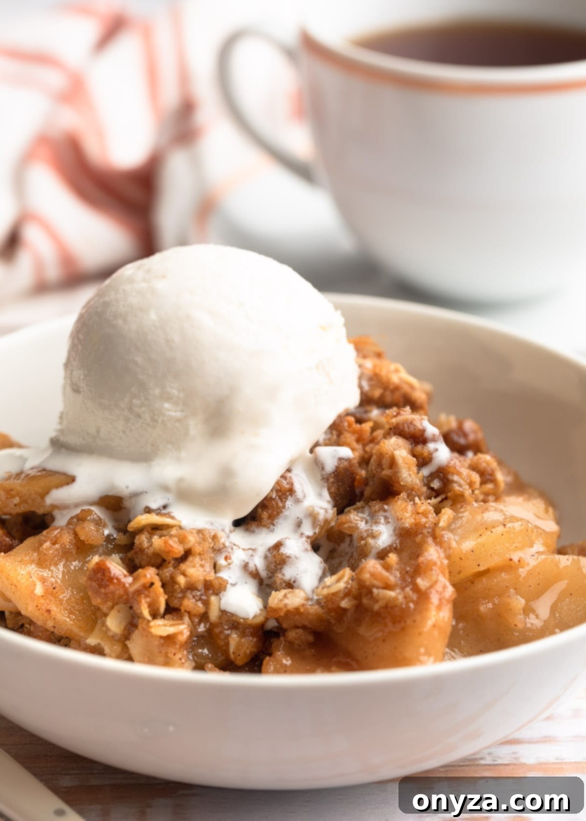 A beautifully presented bowl of homemade Apple Crisp, topped with a generous scoop of melting vanilla ice cream, with a cozy cup of tea blurred in the background, signaling warmth and comfort.