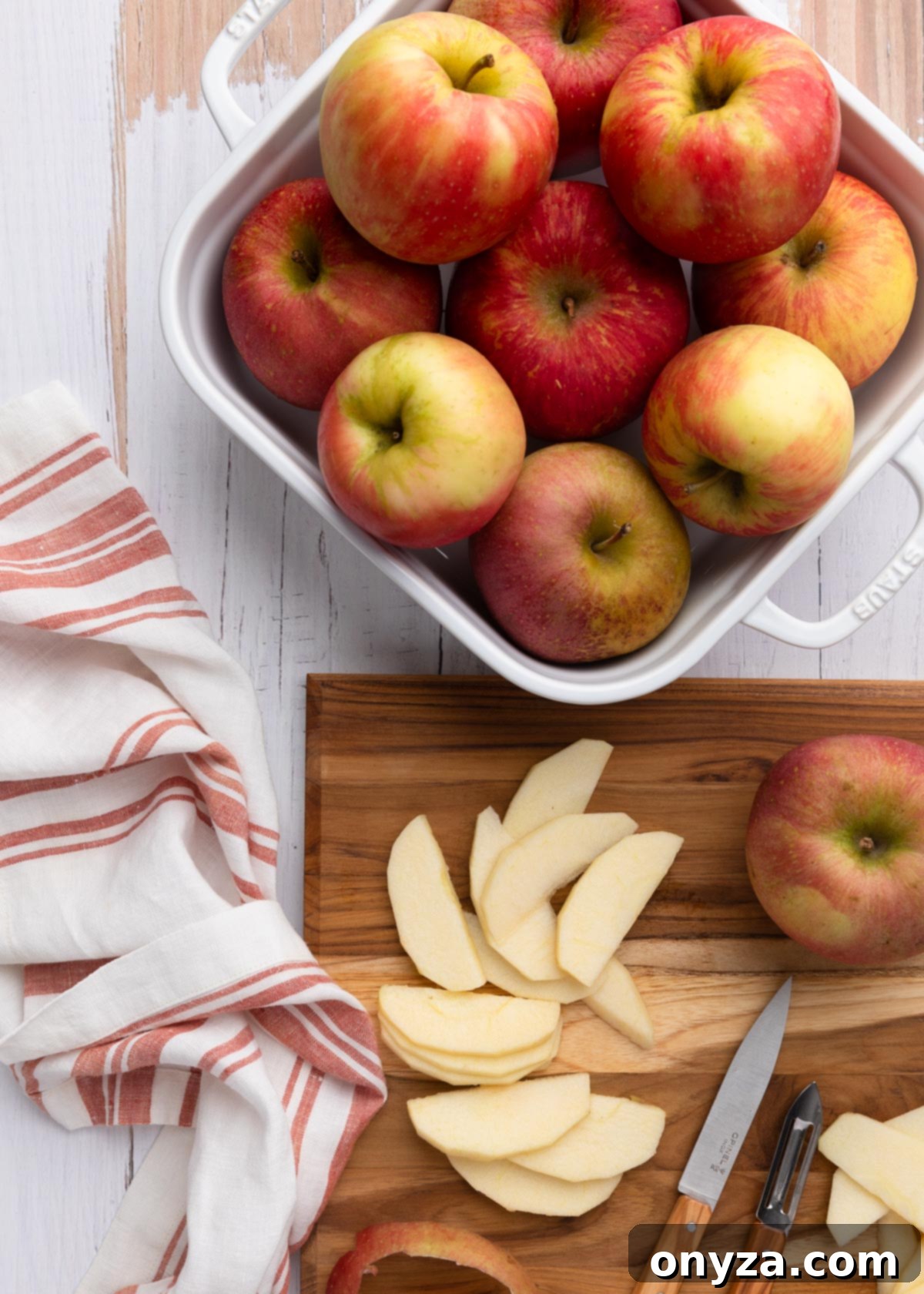 An overhead shot showcasing freshly sliced Honeycrisp apples arranged in a white ceramic baking dish, with a wooden cutting board full of more sliced apples beside it, ready for preparation.