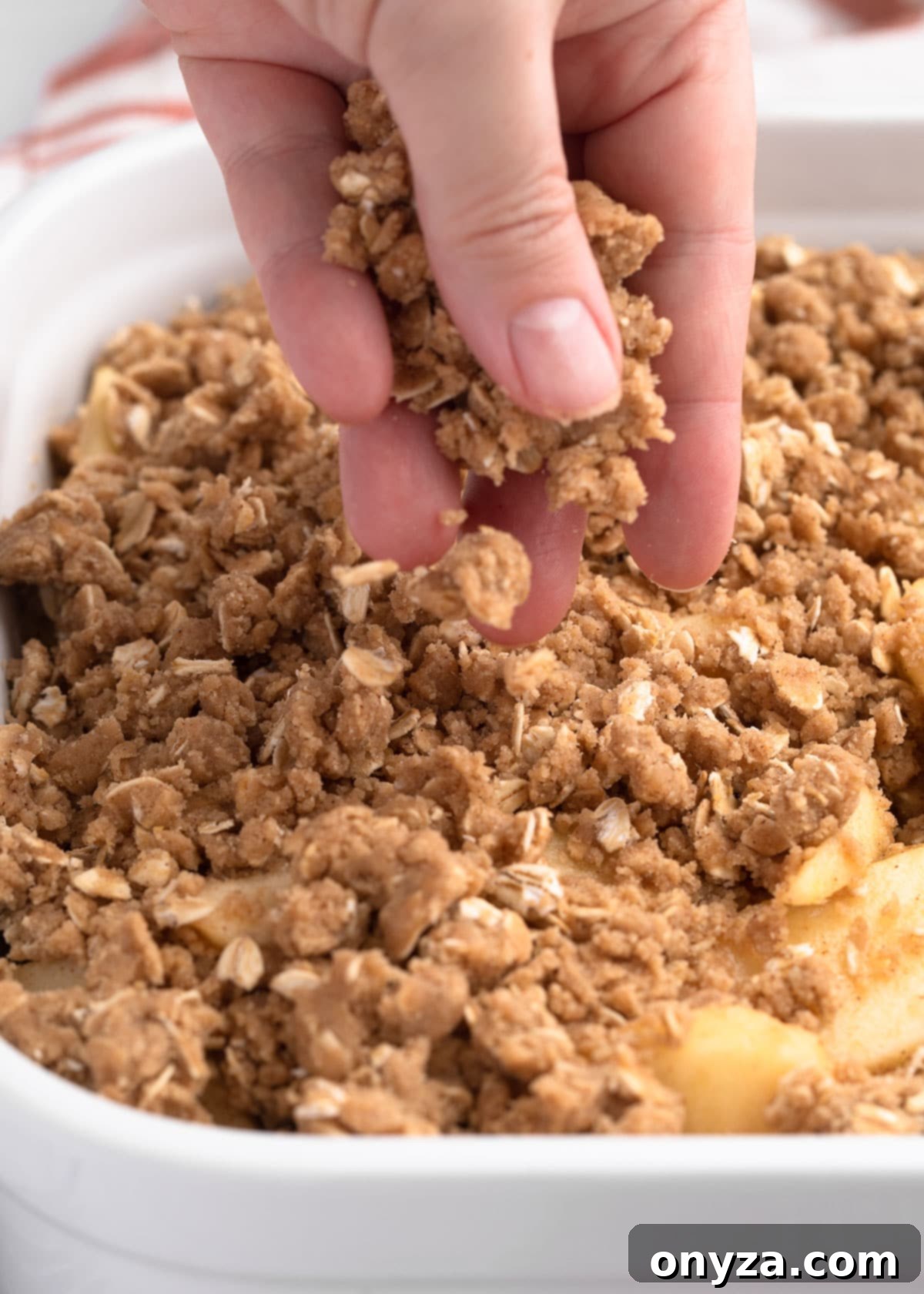 A hand gently sprinkling the golden, crumbly crisp topping evenly over a bed of spiced apple slices in a white ceramic baking dish, ready for the oven.