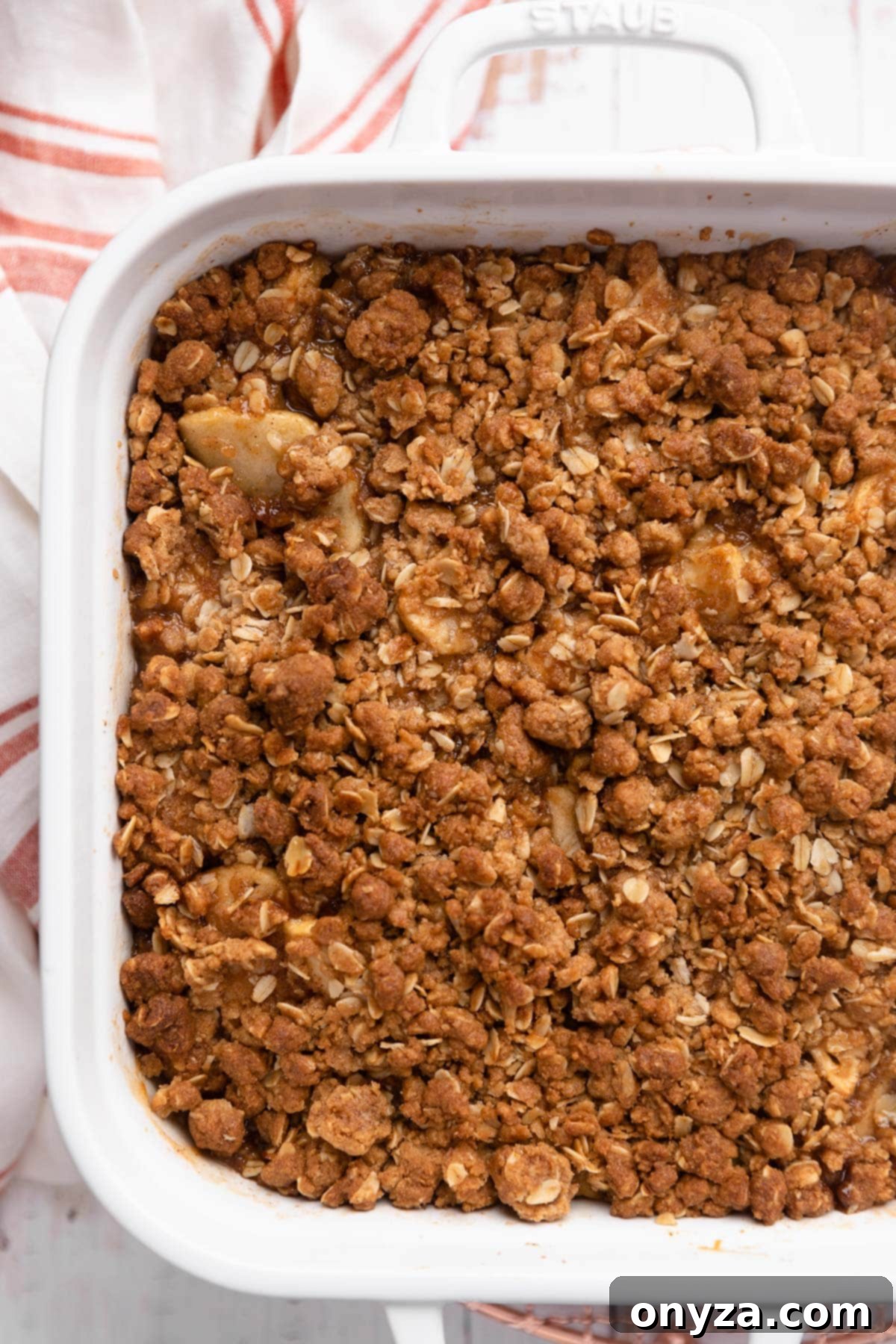 A golden-brown, freshly baked apple crisp still warm in its white ceramic dish, with a folded red striped napkin beside it, ready for serving.