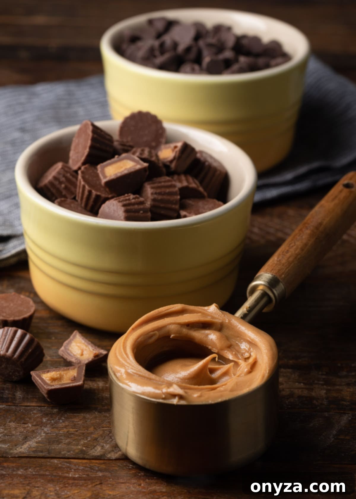 measuring cup filled with creamy peanut butter in front of bowls of mini peanut butter cups and chocolate chips