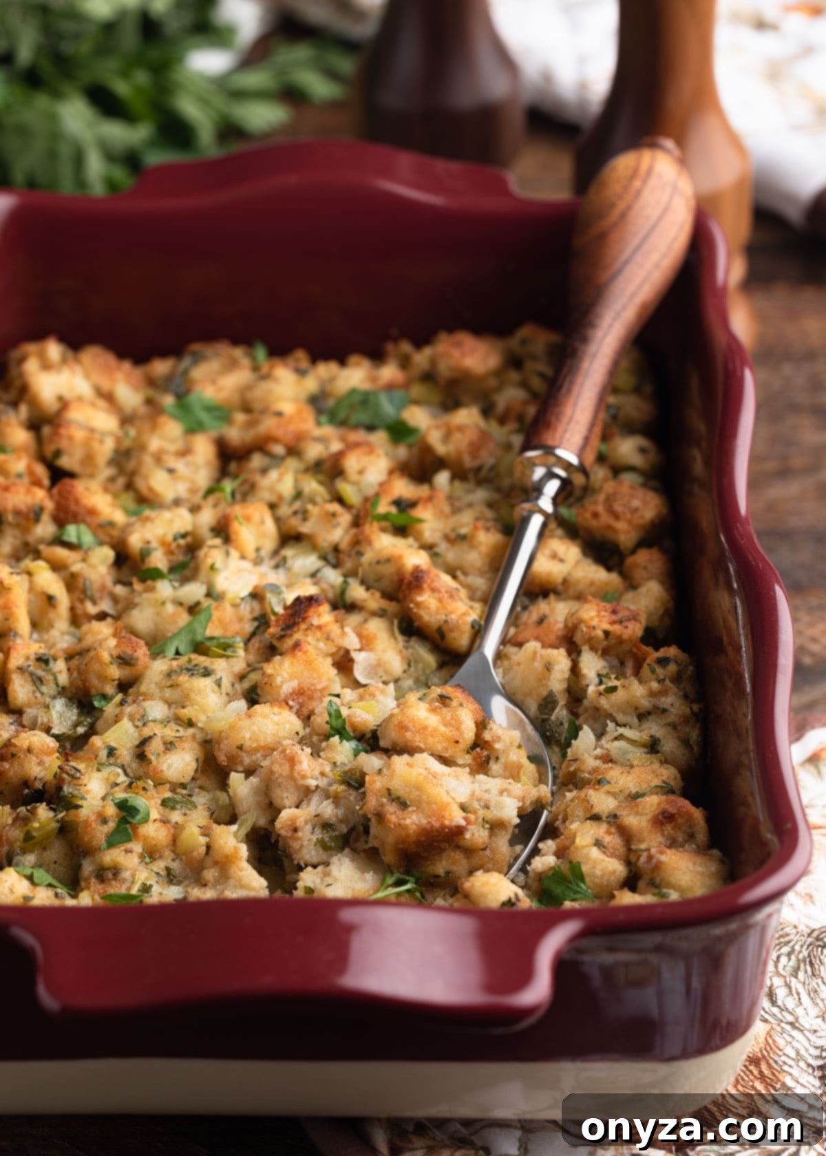 A ceramic baking dish filled with golden-brown, aromatic herb stuffing, with a wood-handled serving spoon resting alongside, ready to be enjoyed.