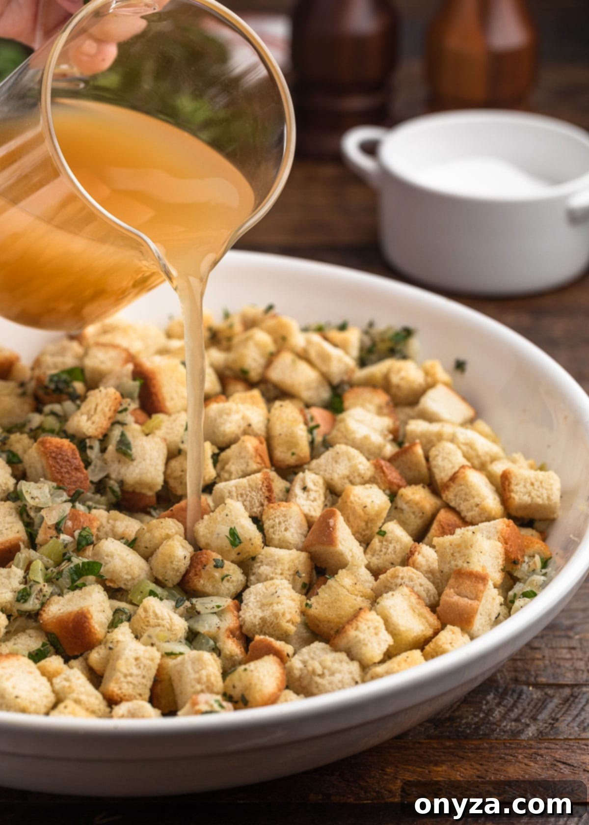 Rich chicken stock being poured over a bowl of dried bread croutons and aromatic vegetables, initiating the soaking process for traditional stuffing.