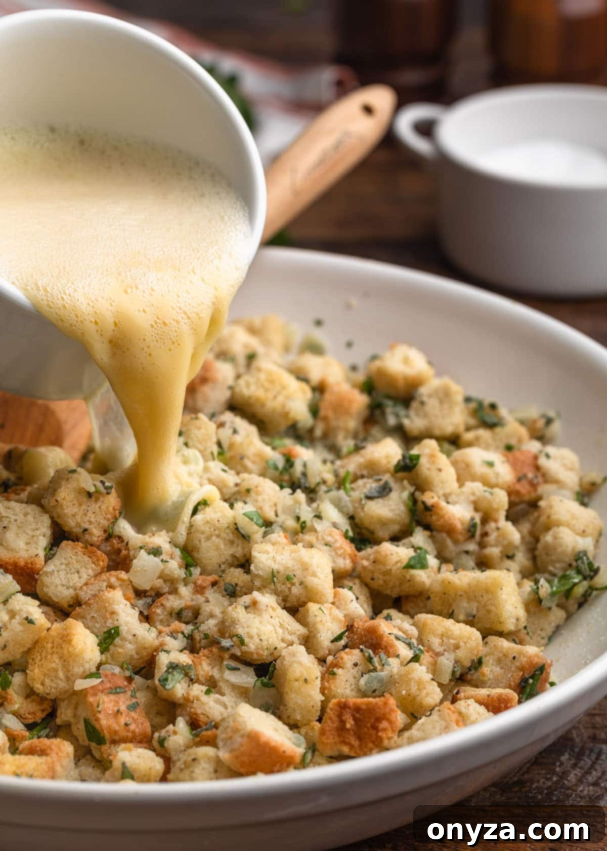 Beaten eggs combined with additional stock being poured into the unbaked bread stuffing mixture in a white bowl, enhancing moisture and binding.