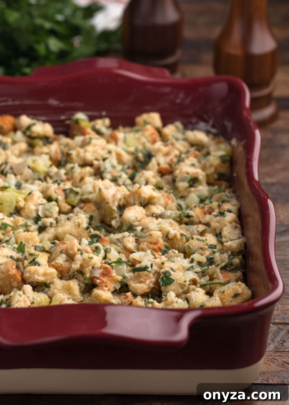 Unbaked herb stuffing meticulously transferred to a burgundy ceramic baking dish, ready for the final touch of butter before being placed in the oven.