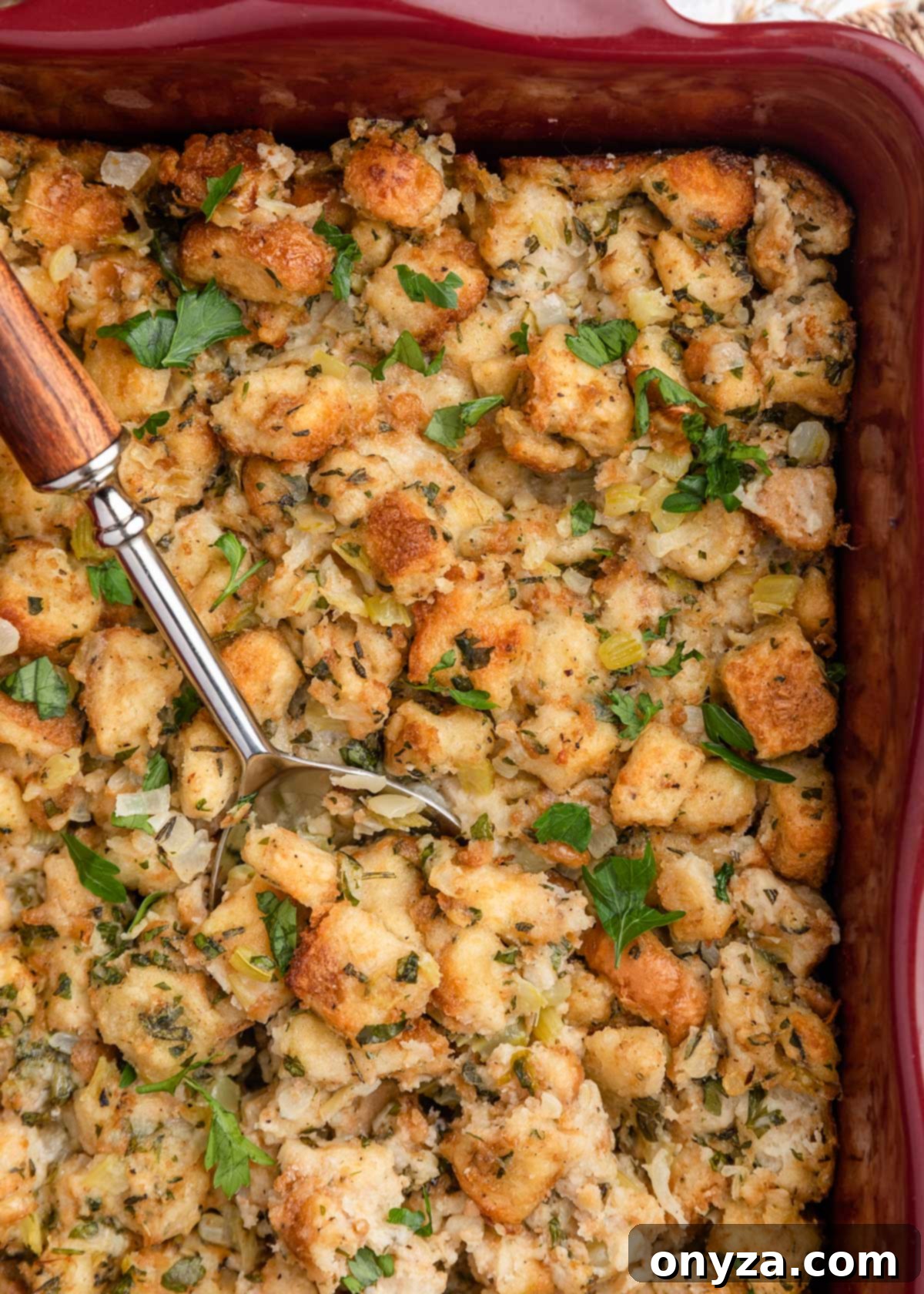 An overhead view of a beautifully baked bread and herb stuffing in a maroon ceramic casserole dish, with a serving spoon nestled within, ready to be enjoyed.