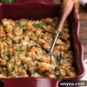 A ceramic baking dish filled with golden-brown, aromatic herb stuffing, with a wood-handled serving spoon resting alongside.