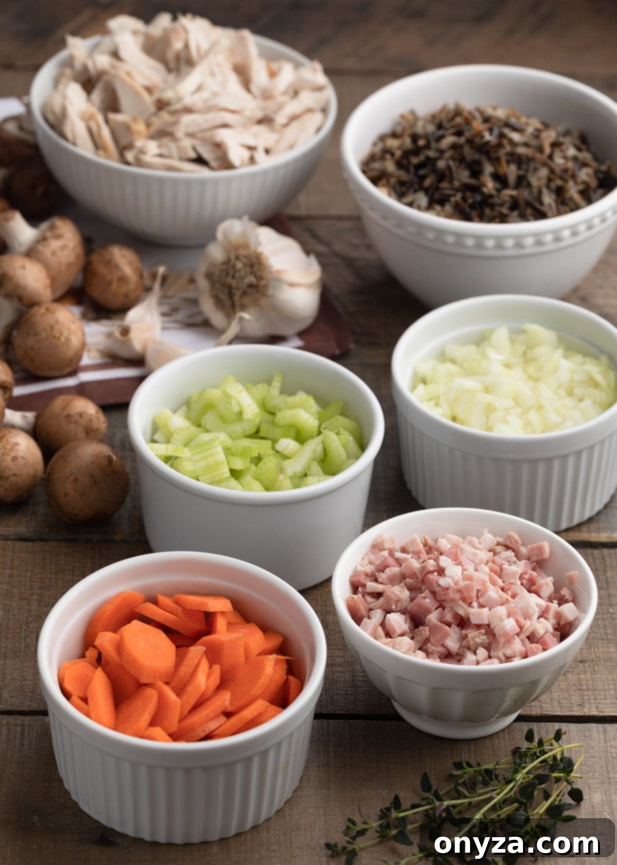 bowls of sliced carrots, pancetta, celery, chopped onions, cooked wild rice, and shredded turkey breast on a wood board, with mushrooms, herbs, and garlic cloves