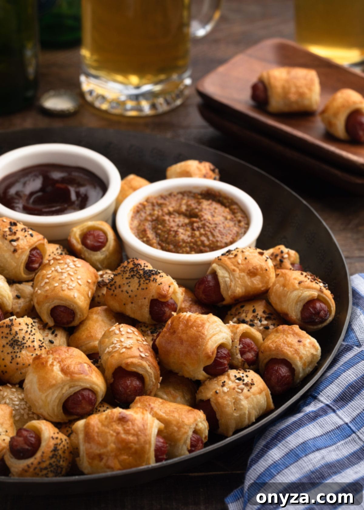 Puff pastry pigs in a blanket on a bronze serving platter with bowls of grainy mustard and bbq sauce for dipping