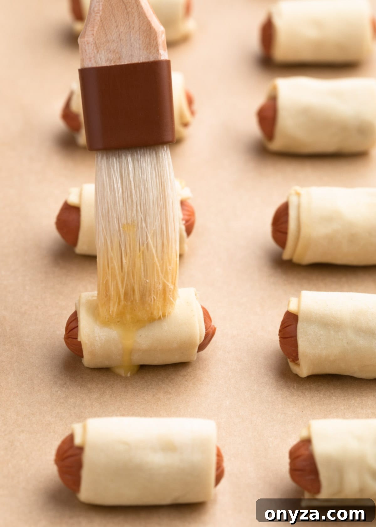 Brushing uncooked pigs in a blanket with egg wash on a baking sheet before baking