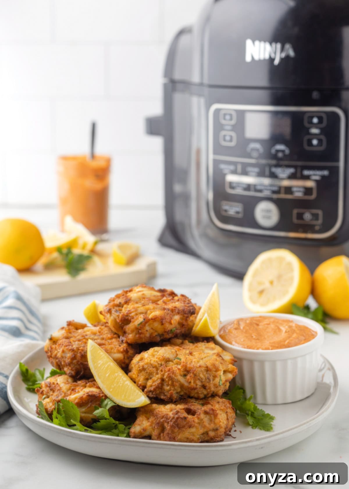Golden brown air fryer crab cakes on a white plate, garnished with fresh lemon wedges and a side of remoulade sauce, with an air fryer in the background.