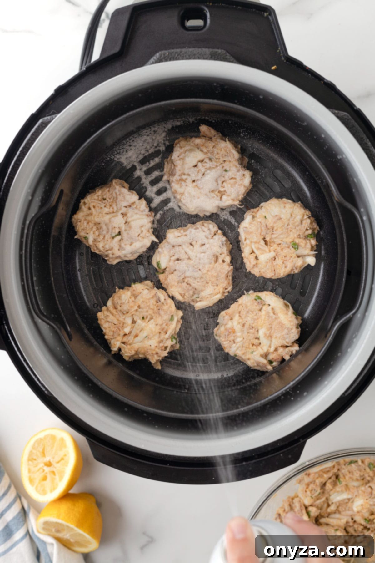 Overhead view of two crab cakes being sprayed with cooking oil inside an air fryer basket.