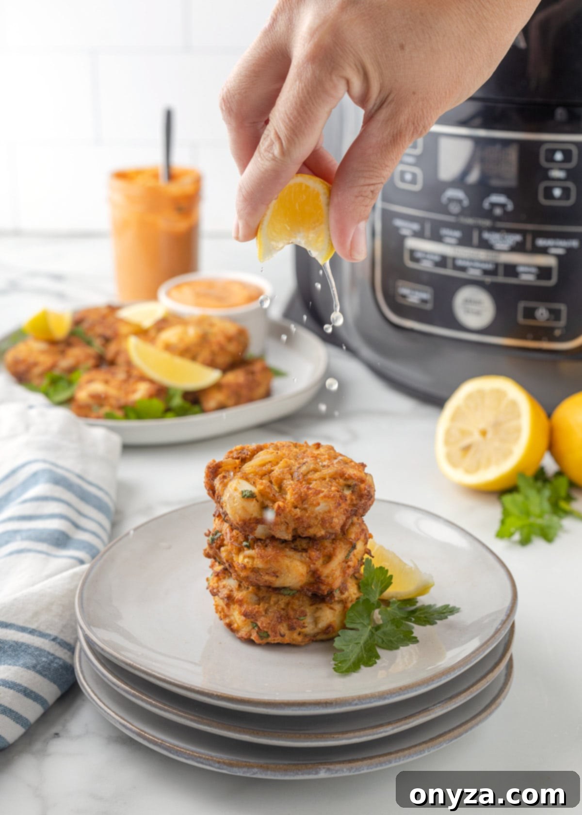 A hand squeezing fresh lemon juice over three stacked air fried crab cakes on gray plates.