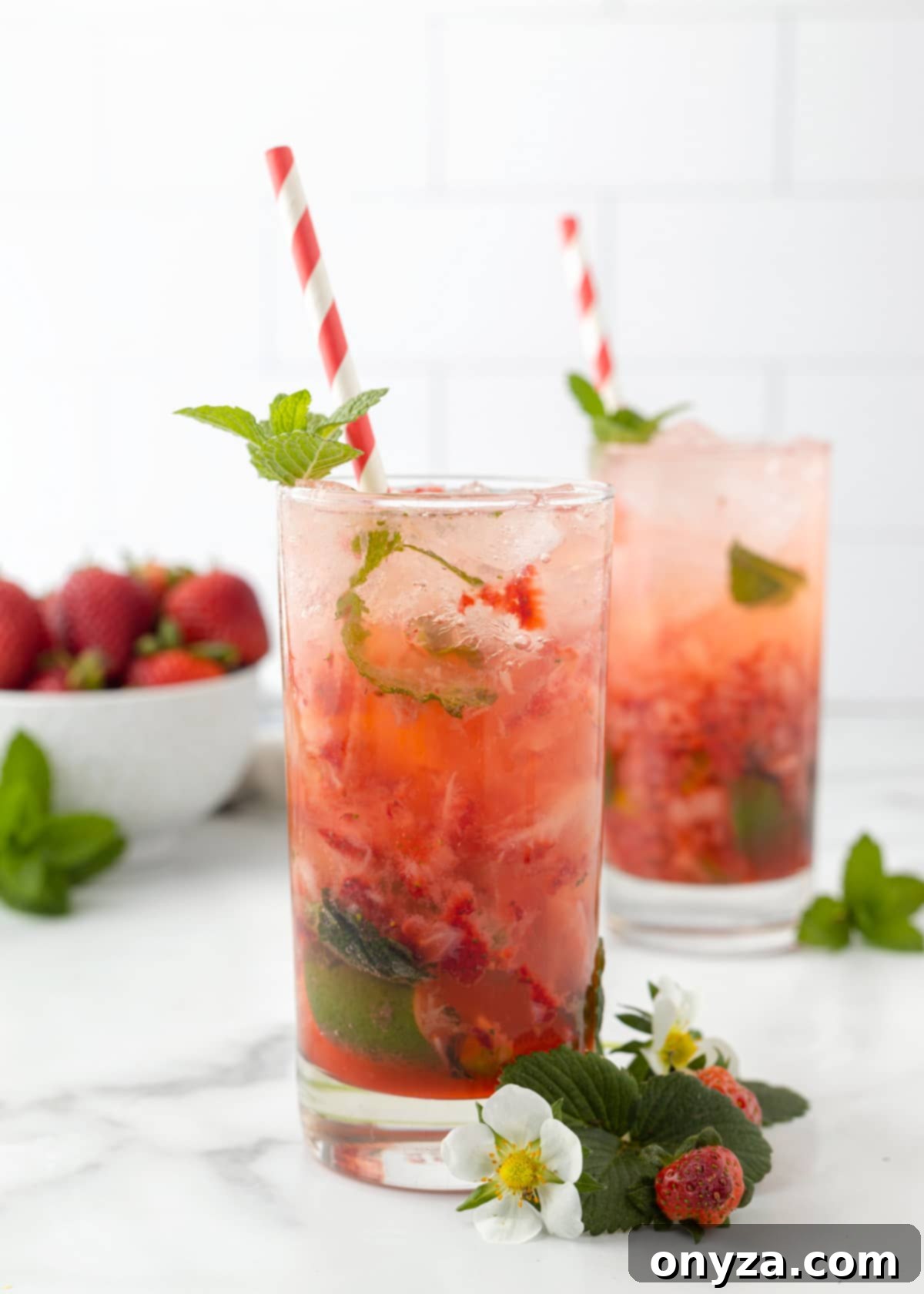 Two vibrant Strawberry Mojitos in highball glasses, garnished with fresh strawberries, mint sprigs, and red and white paper straws, ready to be enjoyed.