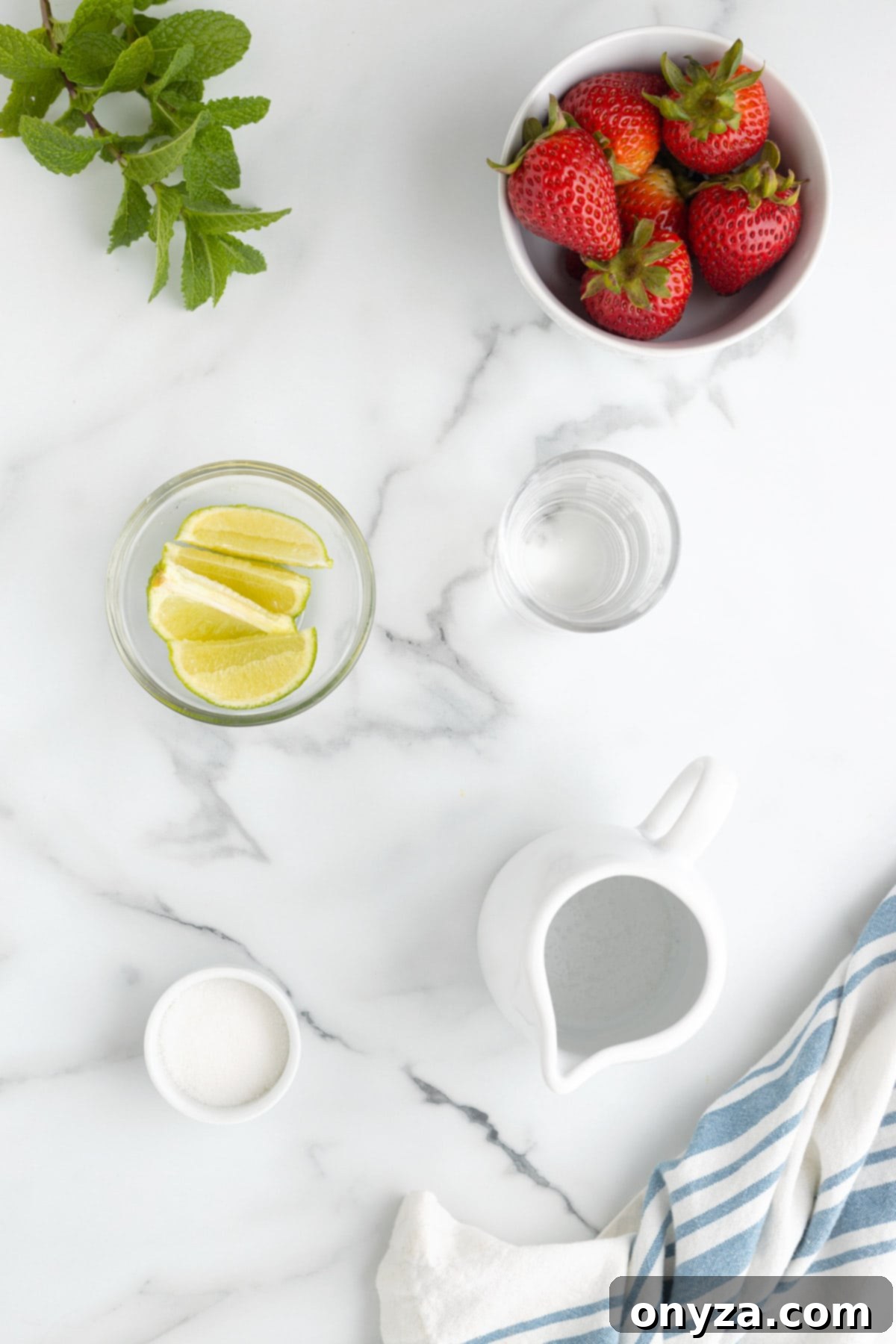 Overhead shot of fresh ingredients for Strawberry Mojito: a bowl of vibrant strawberries, a bowl of bright green lime wedges, a mound of superfine sugar, a bottle of white rum, a fresh sprig of mint, and sparkling water, all arranged on a white marble background.