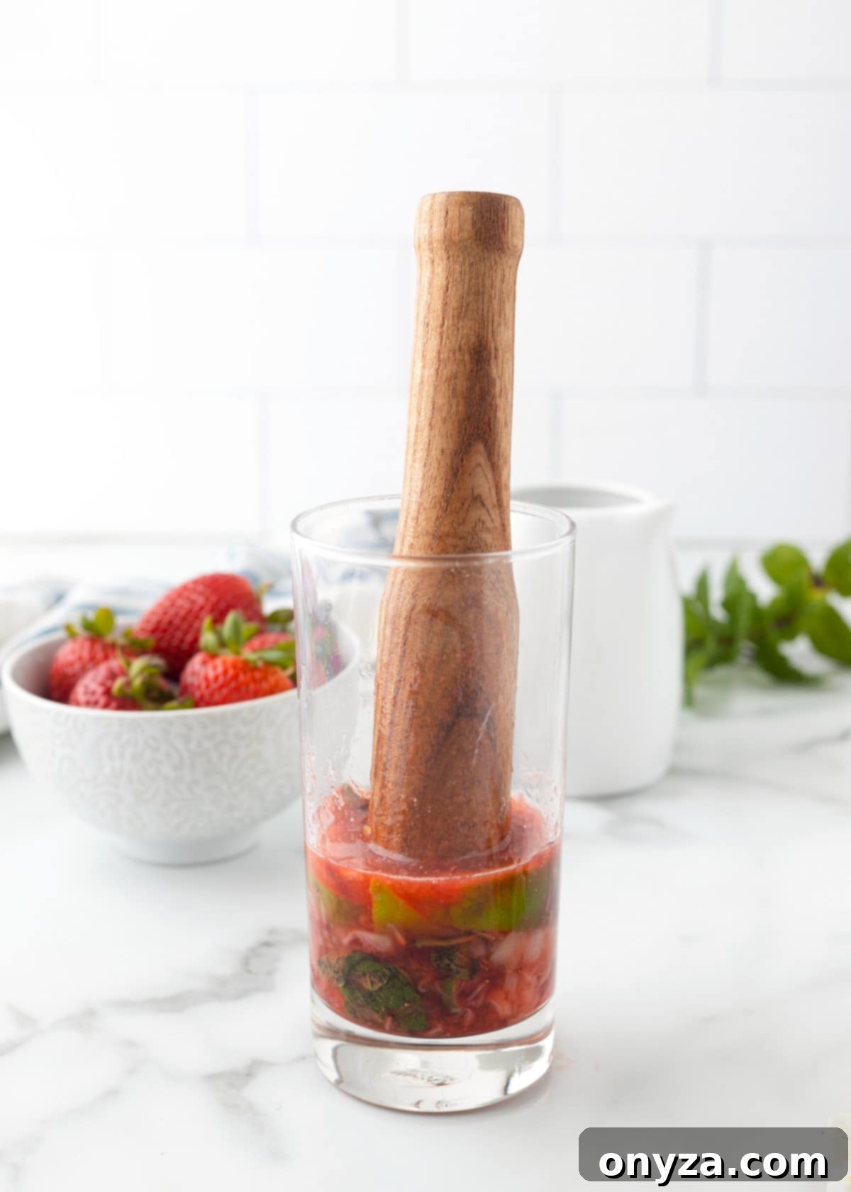 A wooden muddler gently presses down on fresh strawberries, lime wedges, and mint leaves at the bottom of a clear highball glass, illustrating the first step of making a Strawberry Mojito.