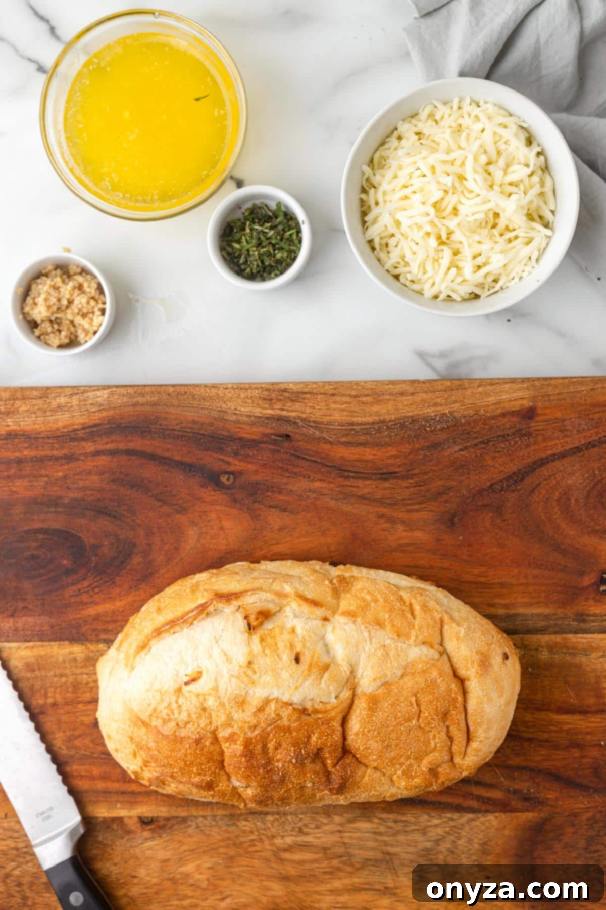 overhead of a loaf of Italian bread on a wood cutting board with bowls of melted butter, shredded mozzarella, chopped herbs, and chopped garlic on white marble, ready for assembly
