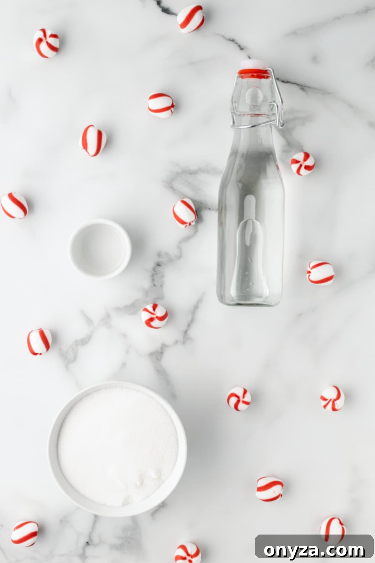 Overhead view of three simple ingredients: a glass bottle of purified water, a bowl of granulated sugar, and a small bowl of pure peppermint extract, arranged on a marble background with scattered peppermint candies.