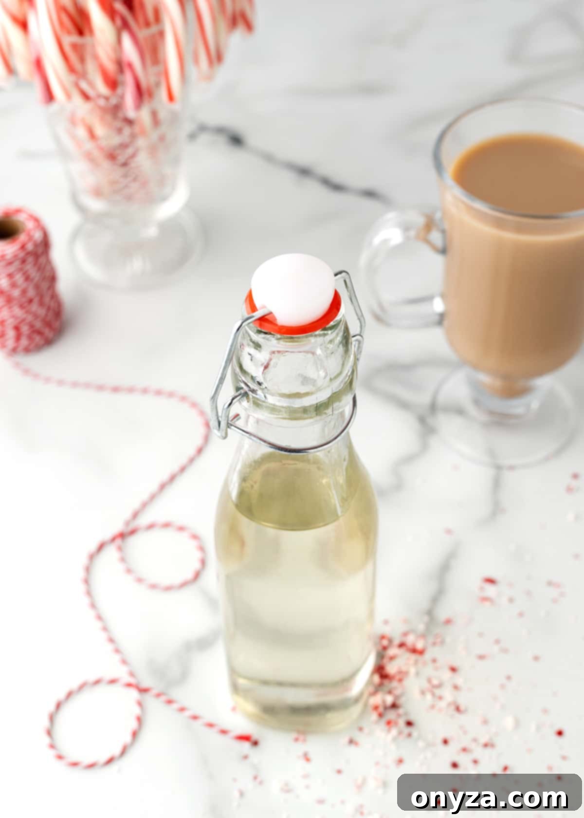 Overhead photo featuring a bottle of homemade peppermint syrup alongside a steaming cup of coffee, candy canes, and festive red baker's string, ready for holiday gifting.