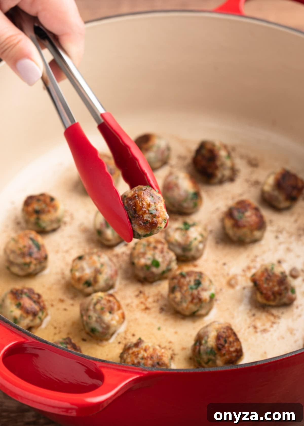 A pair of red tongs carefully removing a seared beef and pork meatball from a hot Dutch oven.