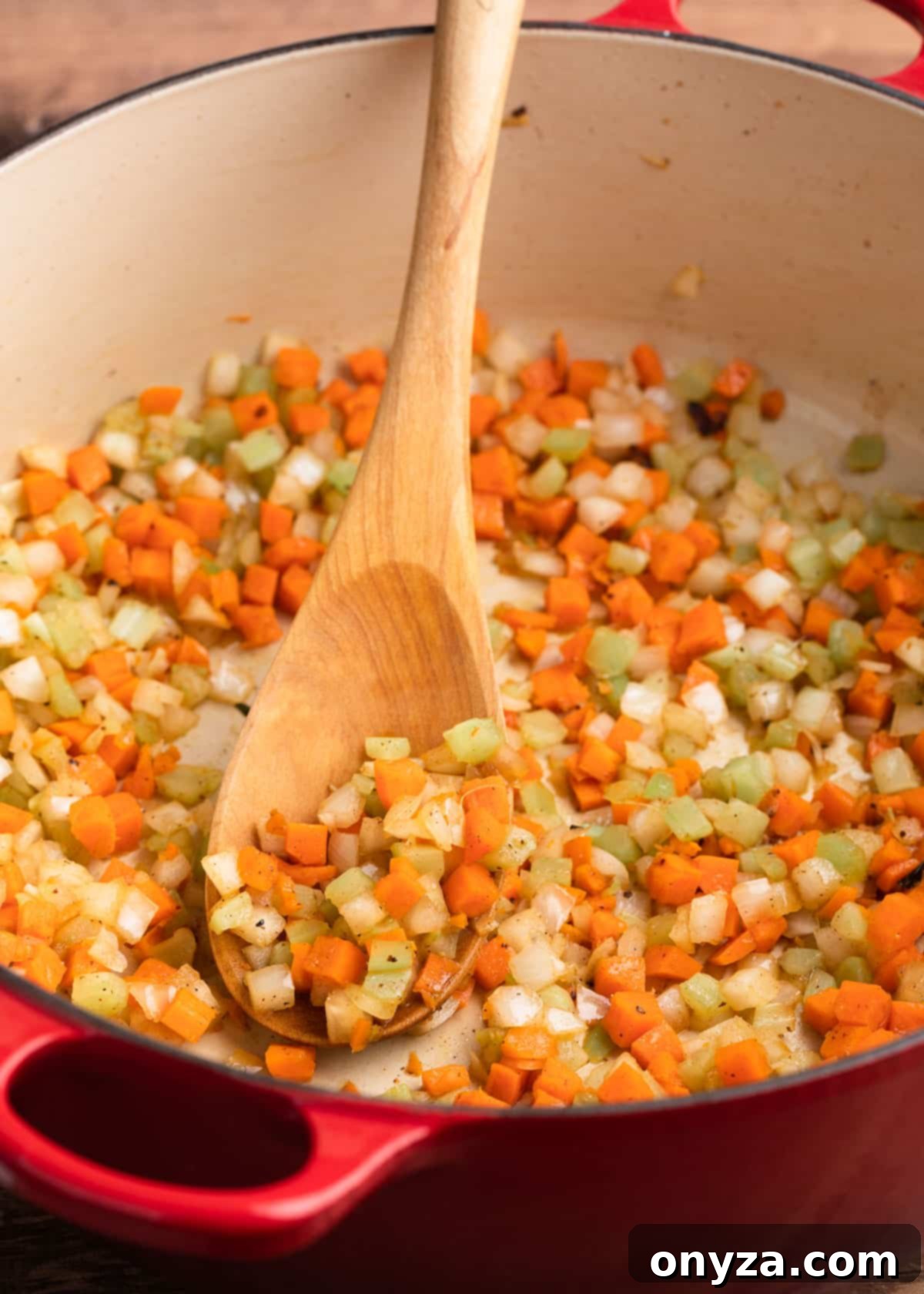 Sautéed celery, carrots, and onions cooking in a red enameled cast iron Dutch oven with a wooden spoon.