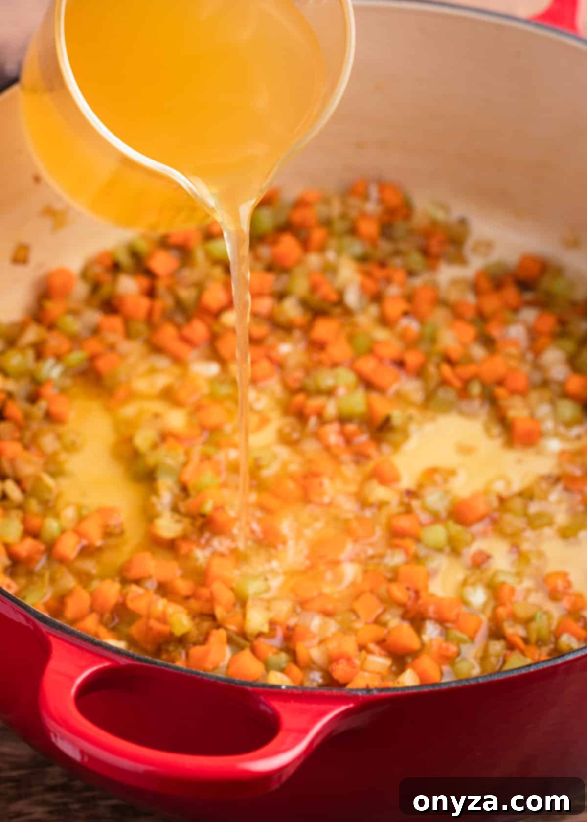 Chicken stock being poured from a glass measuring cup into a red enameled cast iron Dutch oven containing sautéed carrots, celery, and onions.