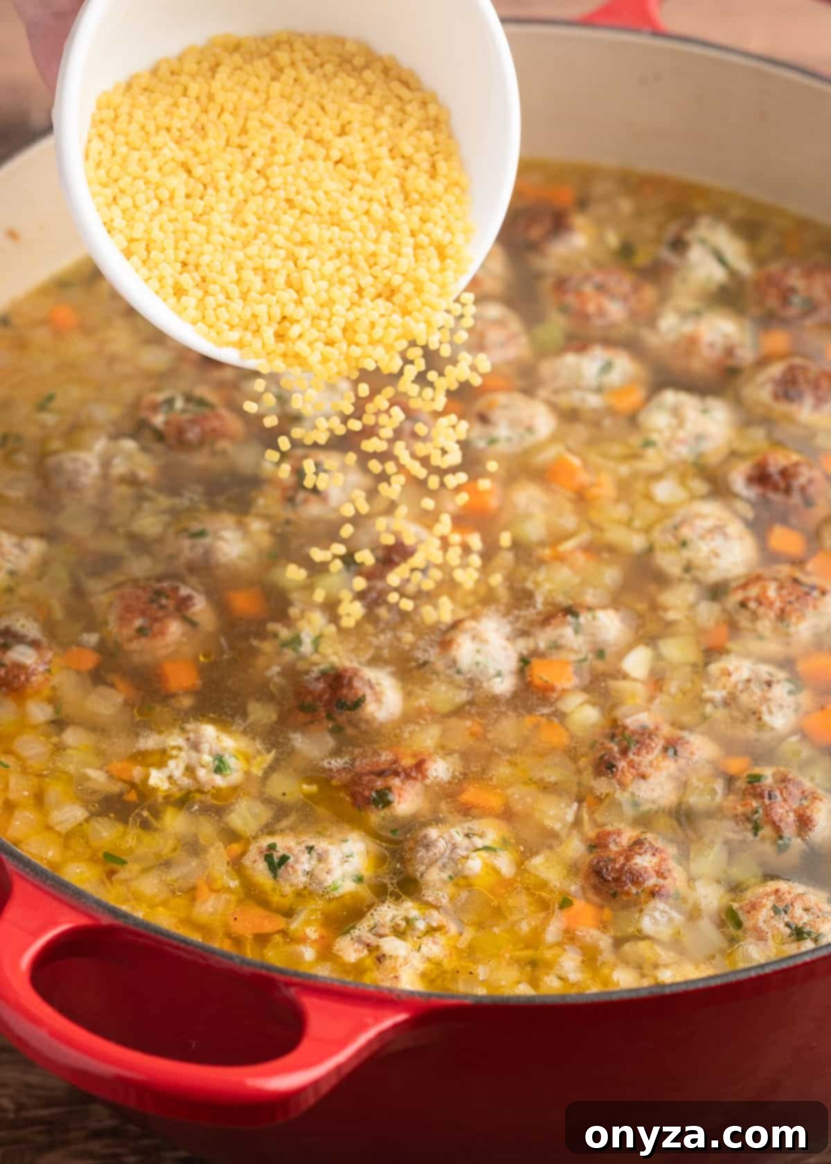 Acini di Pepe pasta being poured from a white ceramic bowl into a bubbling pot of Italian Wedding Soup.