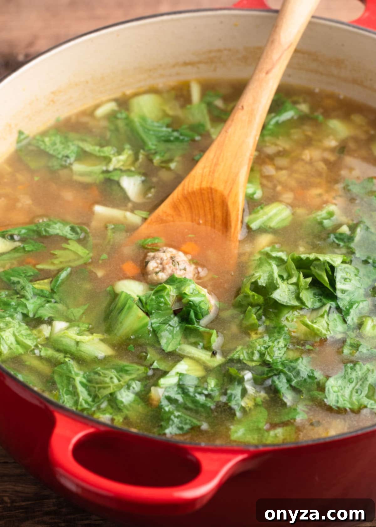 Chopped escarole being stirred into a simmering pot of Italian Wedding Soup with a wooden spoon.