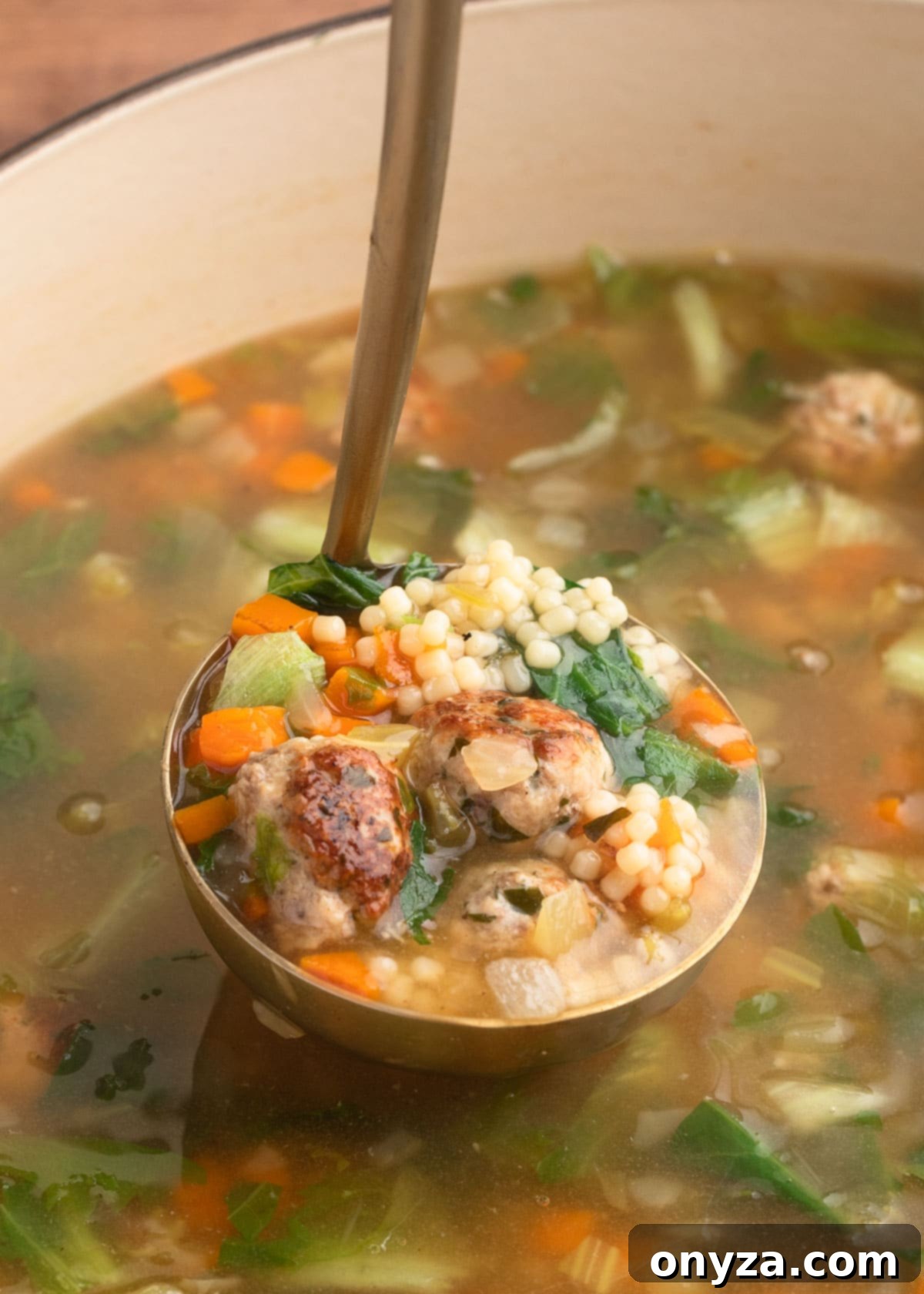 A ladleful of hearty Italian Wedding Soup, brimming with meatballs, pasta, and greens, in an enameled cast iron pot.