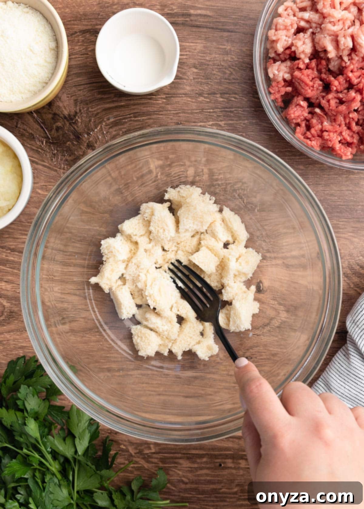 Milk-soaked white bread being mashed with a fork in a glass bowl to create a panade.