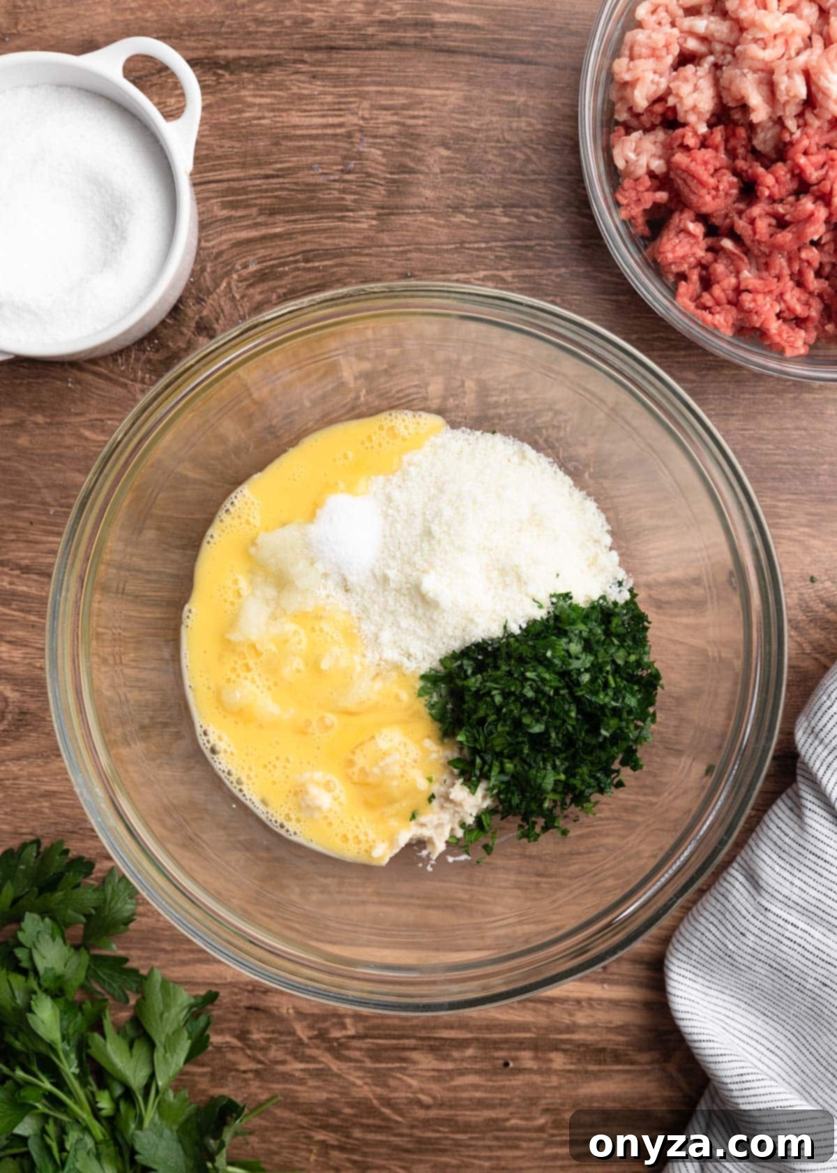 Overhead view of beaten egg, Pecorino Romano cheese, and chopped parsley ready to be mixed into the meatball base.