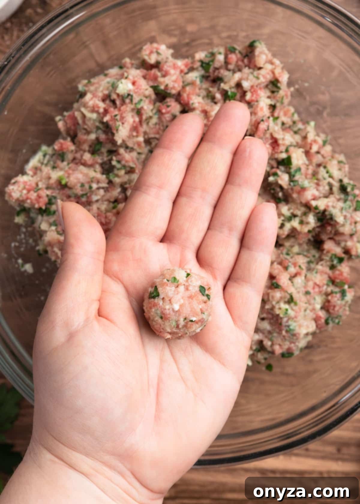 An open hand holding a small, perfectly rolled beef and pork meatball over a bowl of the remaining mixture.