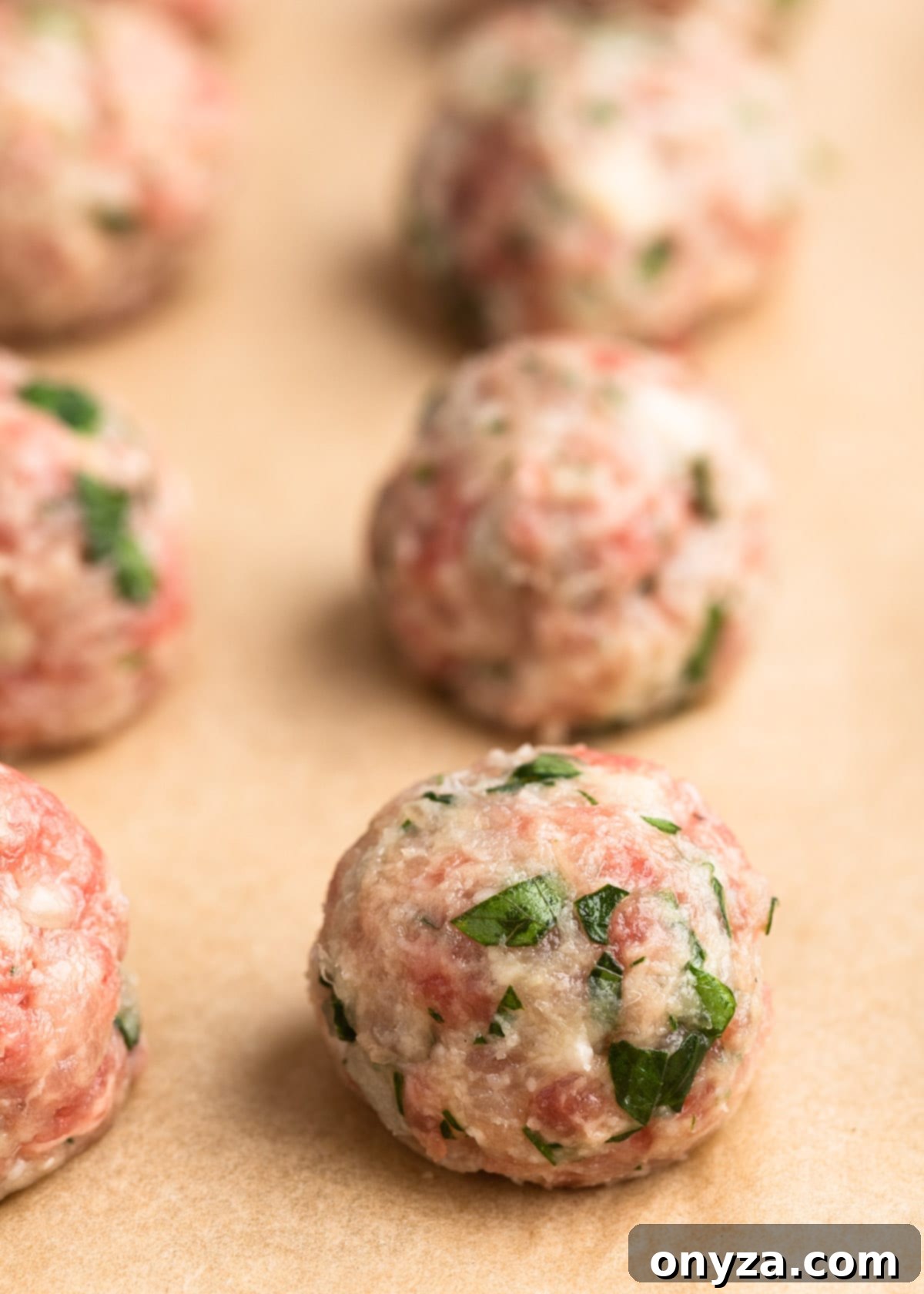 A closeup view of small, uncooked beef and pork meatballs neatly arranged on a parchment-lined baking sheet, ready for browning.