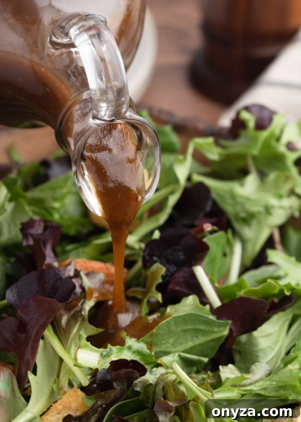 Tangy Balsamic Dressing 5 A close-up shot of rich, golden-brown balsamic vinaigrette being gracefully poured from a clear glass cruet over a vibrant bed of fresh spring mix greens, emphasizing its smooth texture and appetizing sheen.