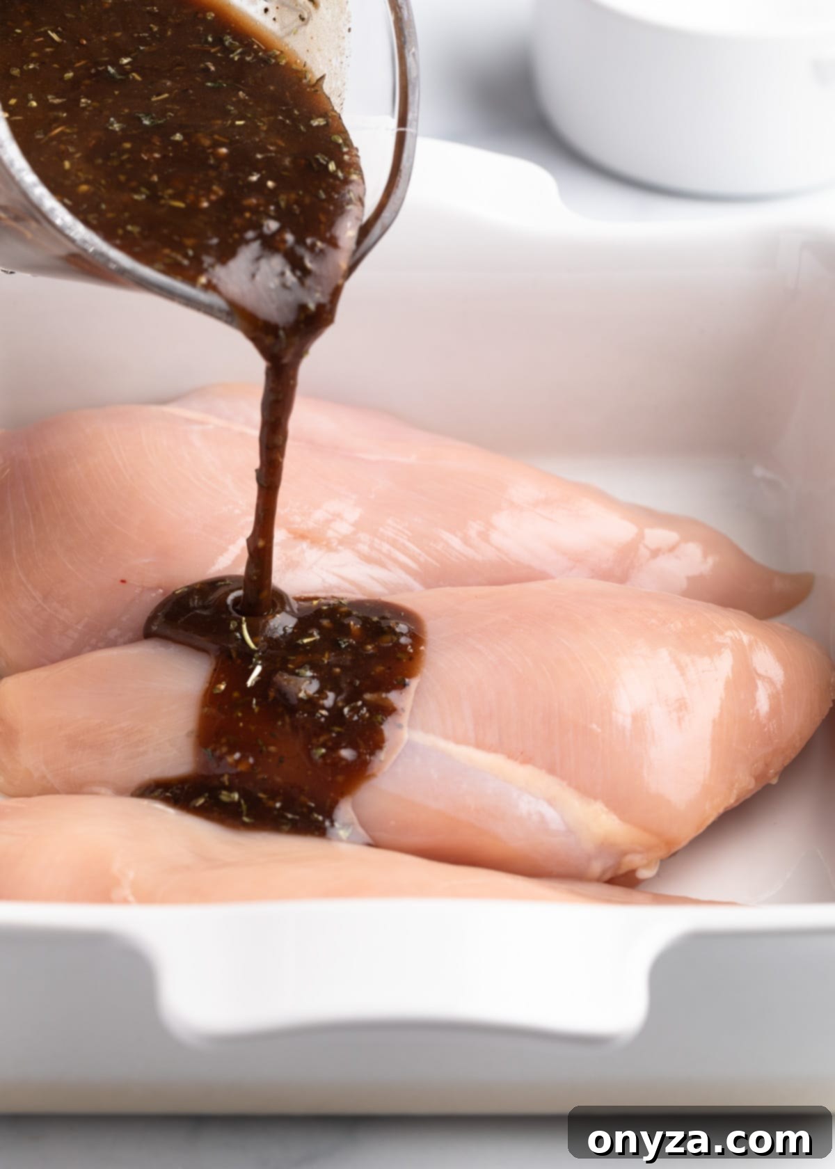 Pouring balsamic marinade over chicken breasts in a white baking dish