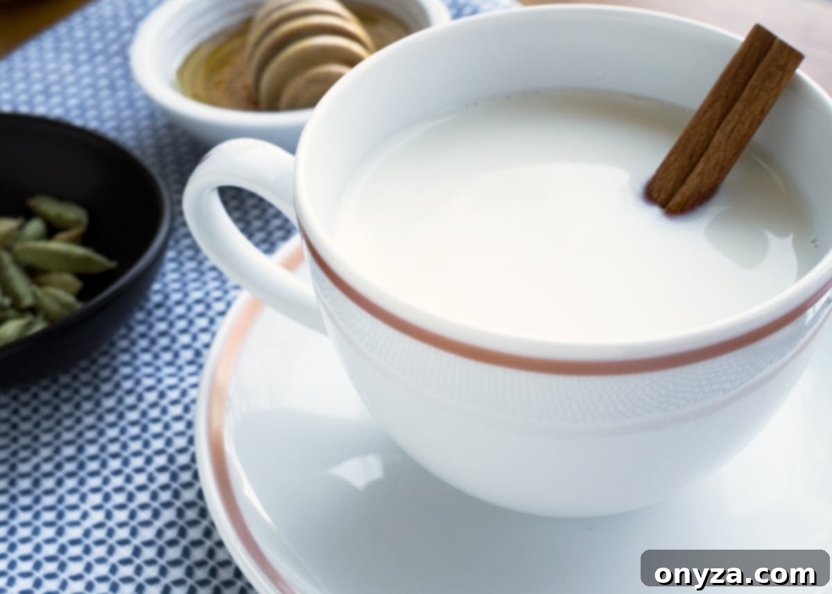 A cozy image of warm cardamom milk served in a white teacup, garnished with a fragrant cinnamon stick, resting invitingly on a blue and white checkered napkin, perfect for a moment of comfort.