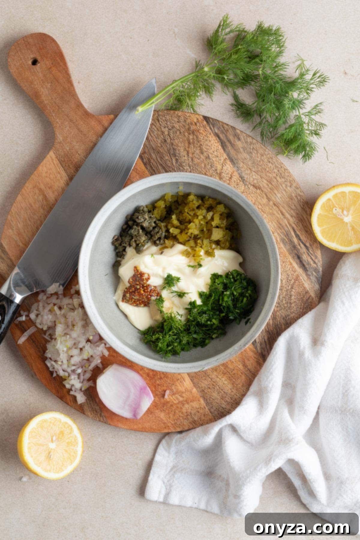 A top-down shot of a rustic wooden cutting board, featuring a pristine white mixing bowl filled with creamy mayonnaise. Around the bowl are carefully arranged small piles of freshly chopped cornichons, aromatic minced shallots, a vibrant mix of fresh herbs (parsley and dill), and briny capers, all prepped and ready to be combined for the tangy tartar sauce.