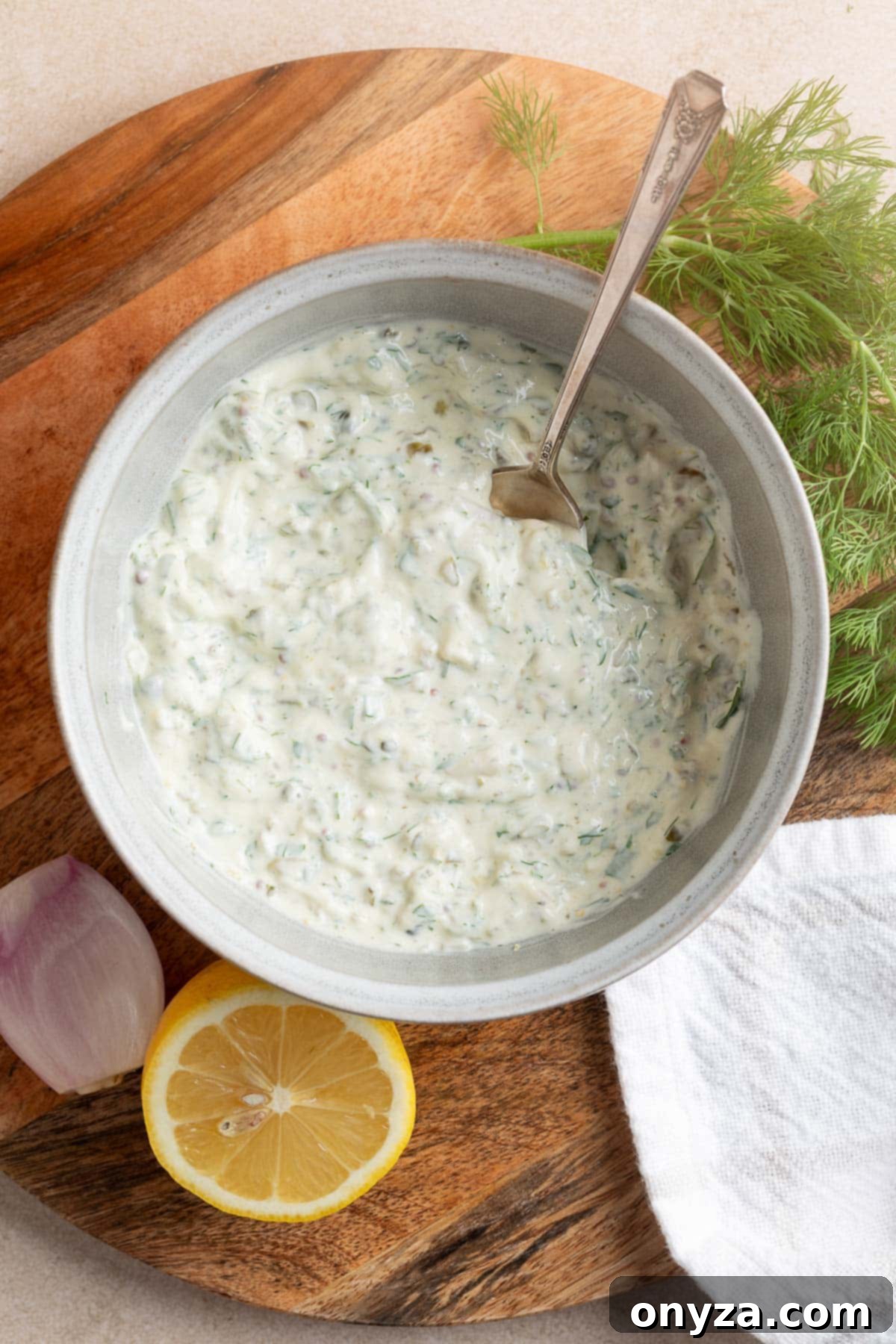 A beautifully mixed bowl of homemade tangy tartar sauce, rich and creamy, presented on a rustic wooden board. A spoon is placed in the bowl, ready to serve, highlighting the sauce's inviting texture and vibrant ingredients.