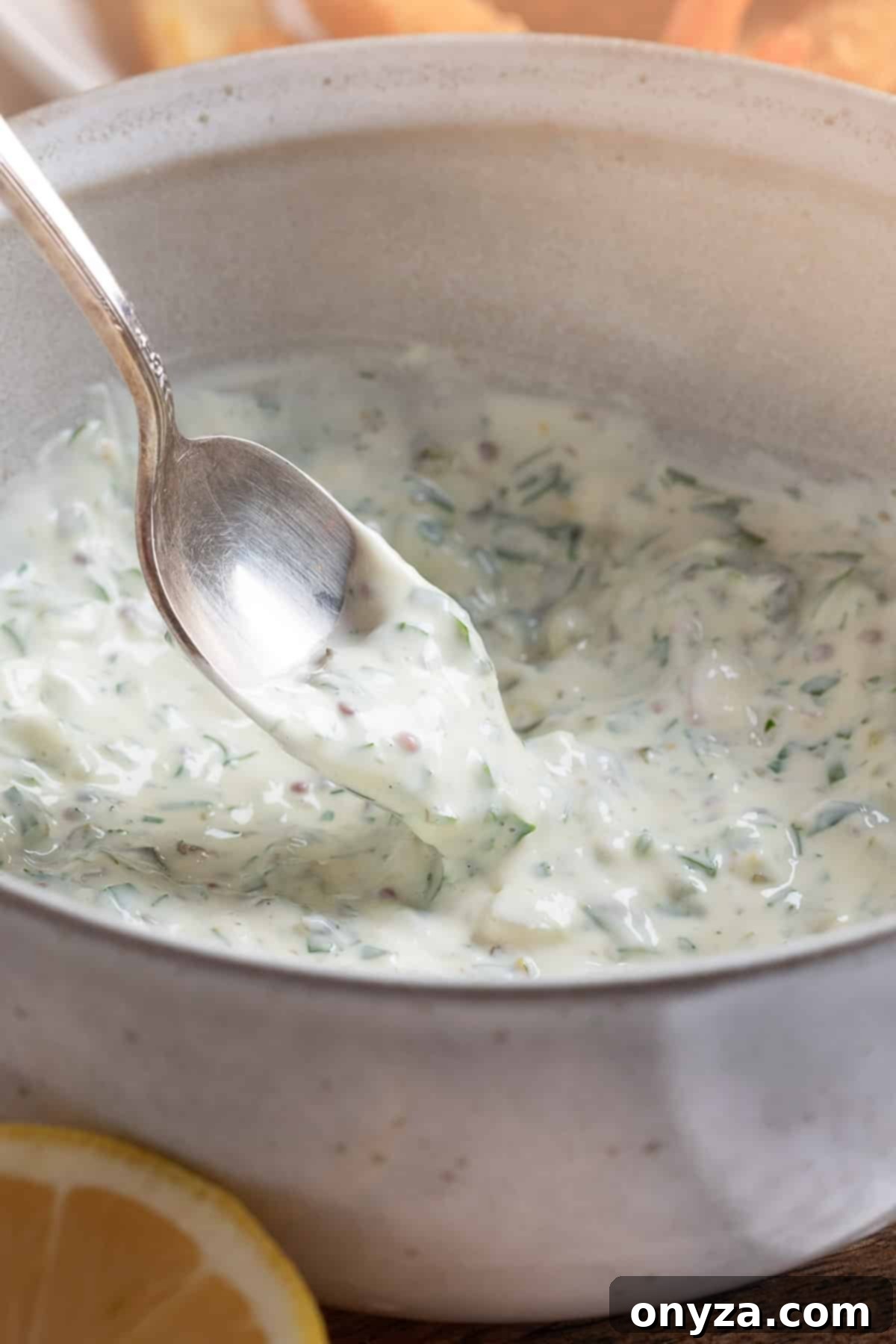 A silver spoon gracefully scoops a generous portion of freshly prepared Tangy Tartar Sauce from a pristine white mixing bowl, showcasing its creamy texture and visible flecks of herbs and cornichons.