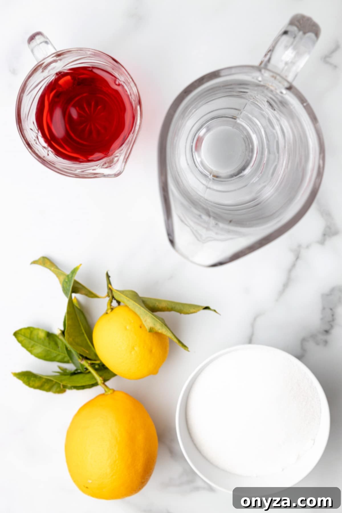 Overhead view of a pristine kitchen counter with clear glass pitchers holding water and cranberry juice, accompanied by fresh lemons and a bowl of granulated sugar, all essential for making homemade pink lemonade.