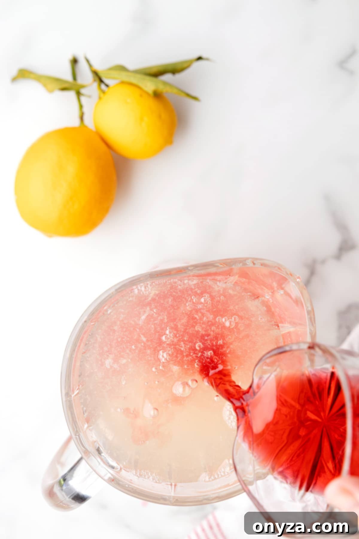 A close-up of cranberry juice being poured into a large glass pitcher filled with fresh lemonade, creating a beautiful pink hue.
