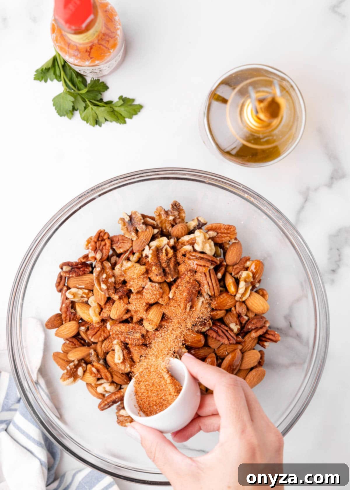 An overhead shot depicting Cajun seasoning being sprinkled into a clear glass bowl filled with mixed nuts, sitting on a pristine marble board, illustrating the seasoning step.