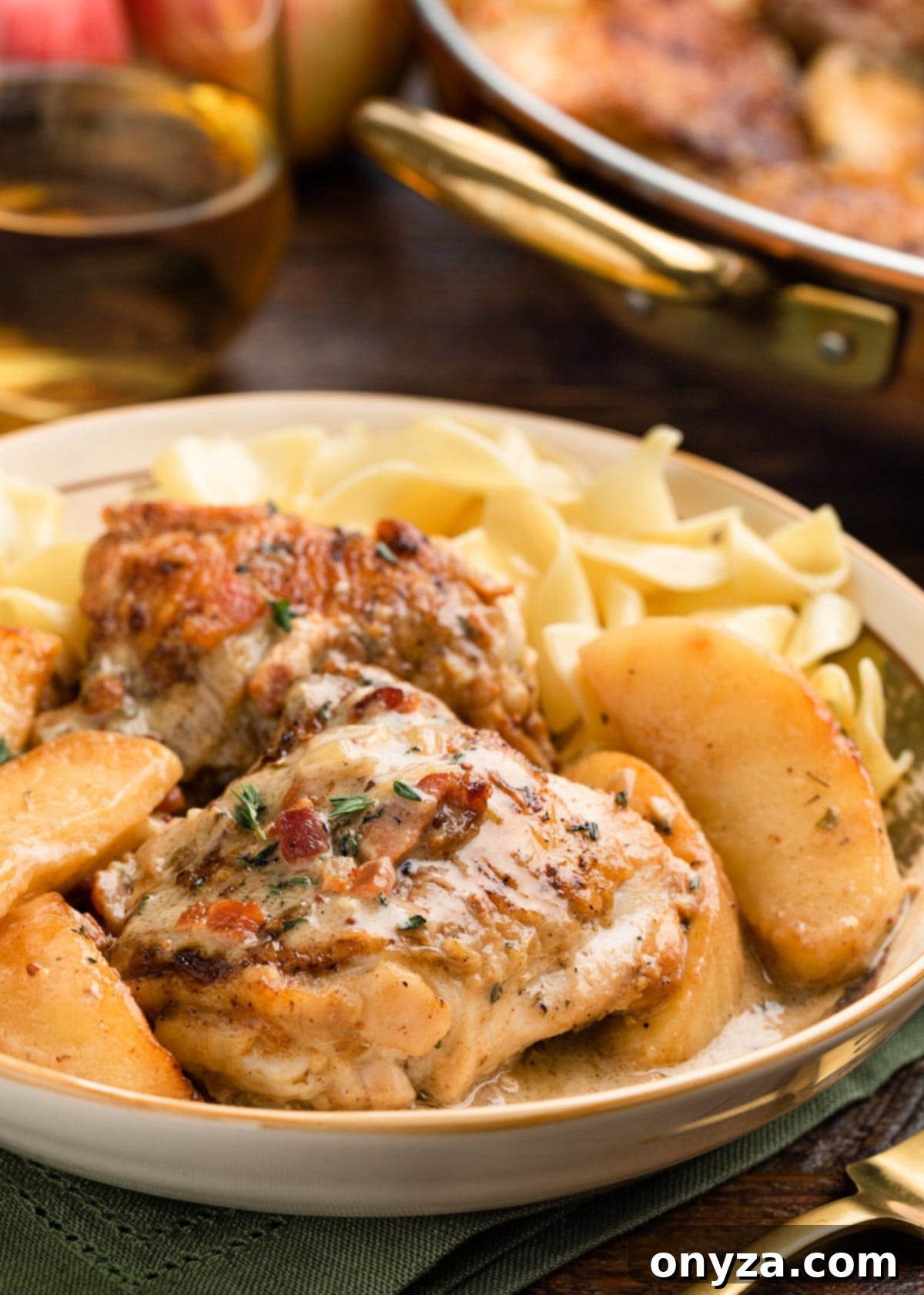 Cider-Braised Chicken with Crispy Bacon and Apples 7 A serving bowl filled with creamy hard cider chicken, tender apples, and sauce, resting on a bed of egg noodles, garnished with fresh herbs – a perfect autumn meal.