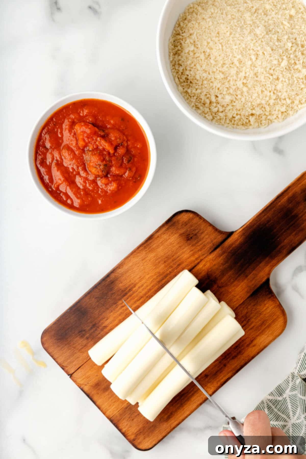 Overhead view of a stack of mozzarella cheese sticks being precisely cut in half crosswise with a chef's knife on a sturdy wood cutting board.