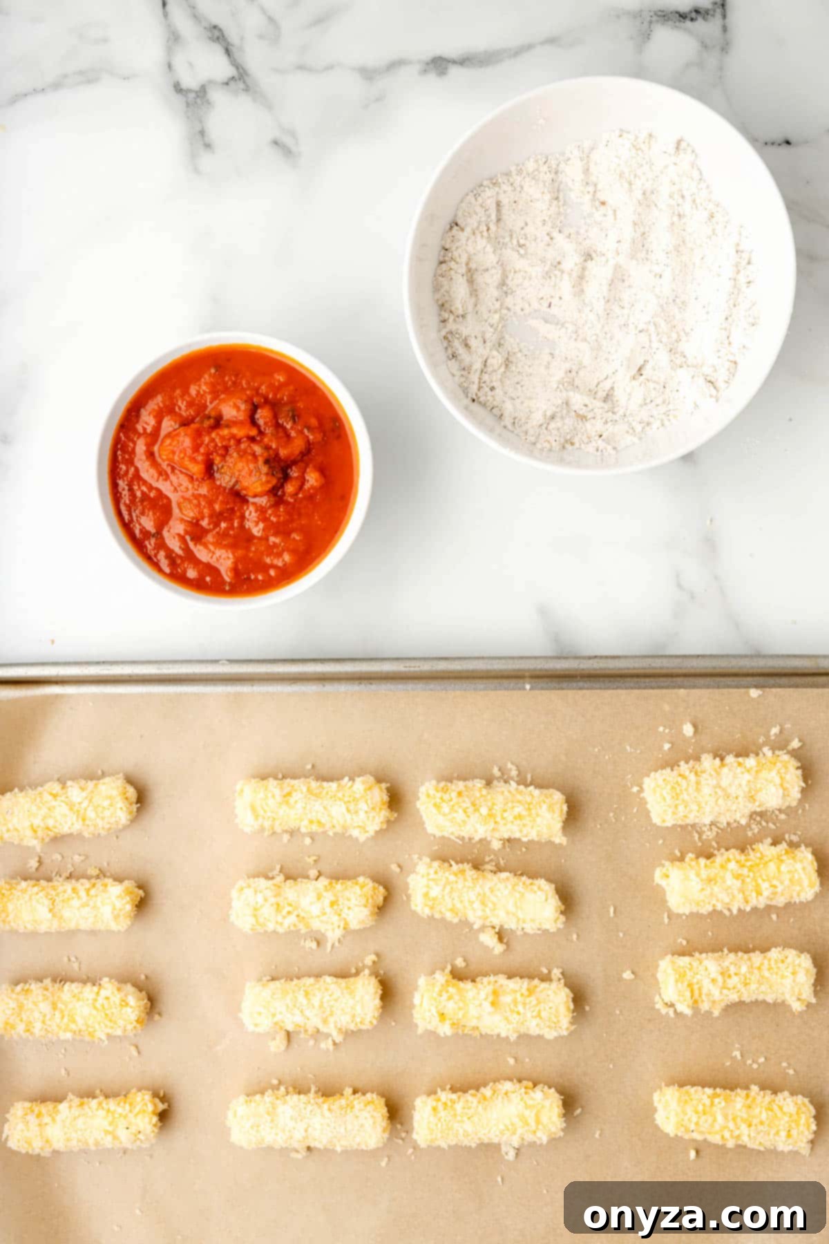 Overhead photo of breaded mozzarella sticks meticulously arranged on a parchment-lined baking sheet, alongside bowls of marinara sauce and flour, ready for freezing.