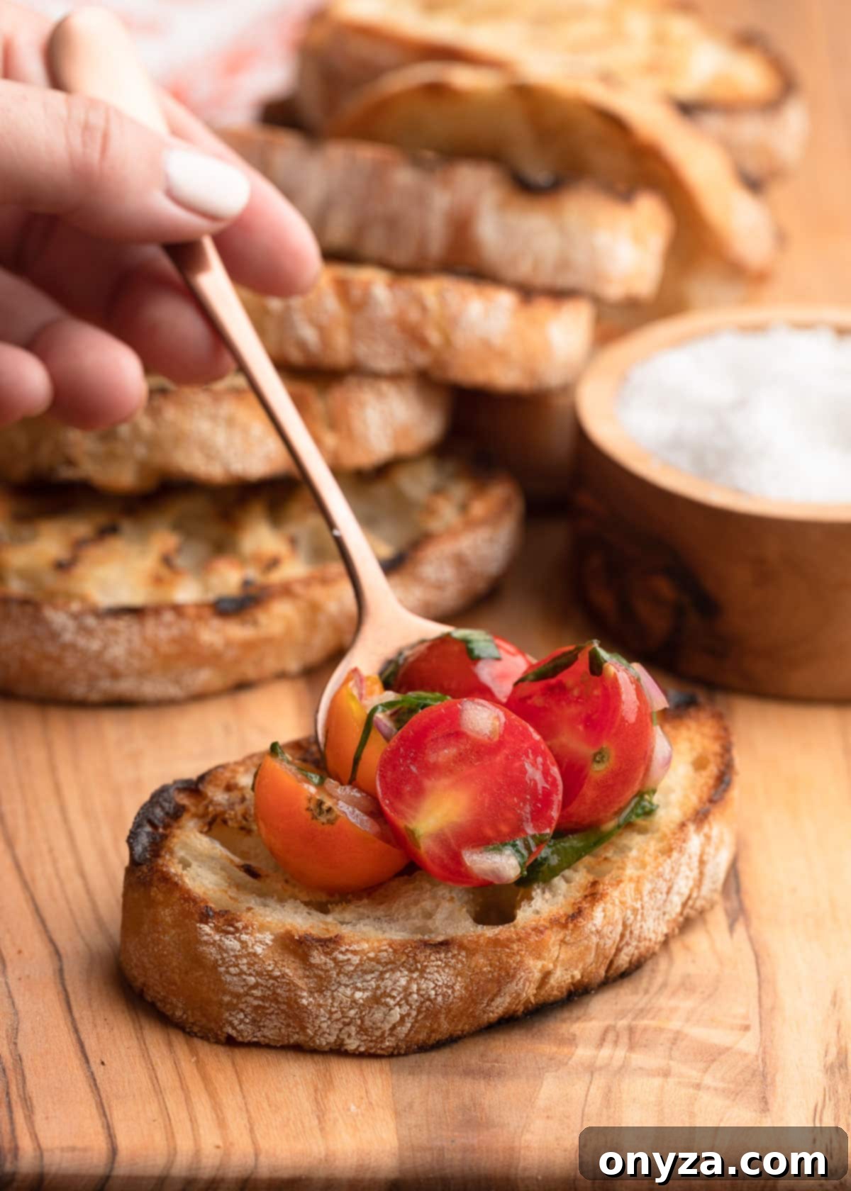 Fresh Cherry Tomato Bruschetta 9 placing cherry tomato bruschetta topping onto a slice of grilled ciabatta bread with a bronze spoon on a wooden board