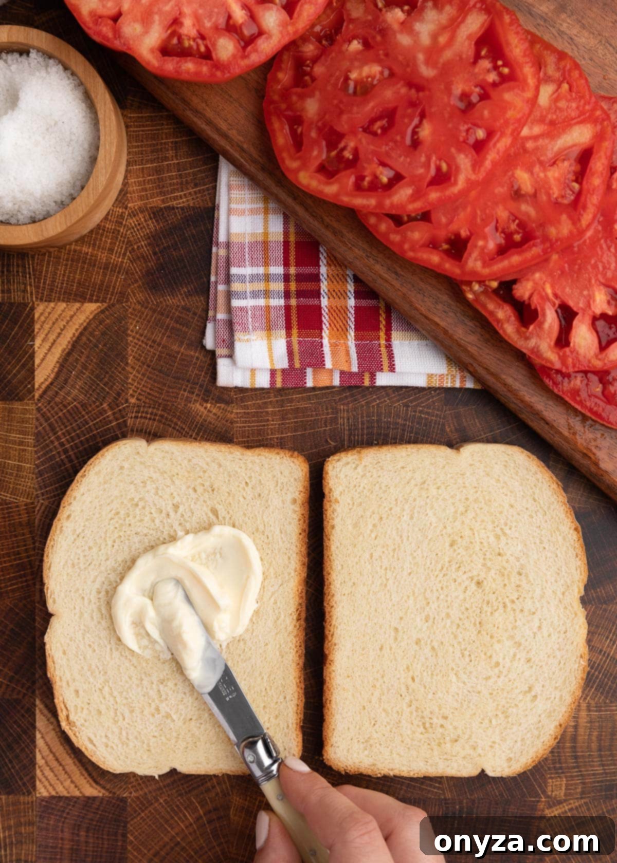 Sun-Kissed Southern Tomato Sandwich 5 Overhead closeup of two slices of white bread on a butcher block board. A knife is spreading mayonnaise onto the left slice.