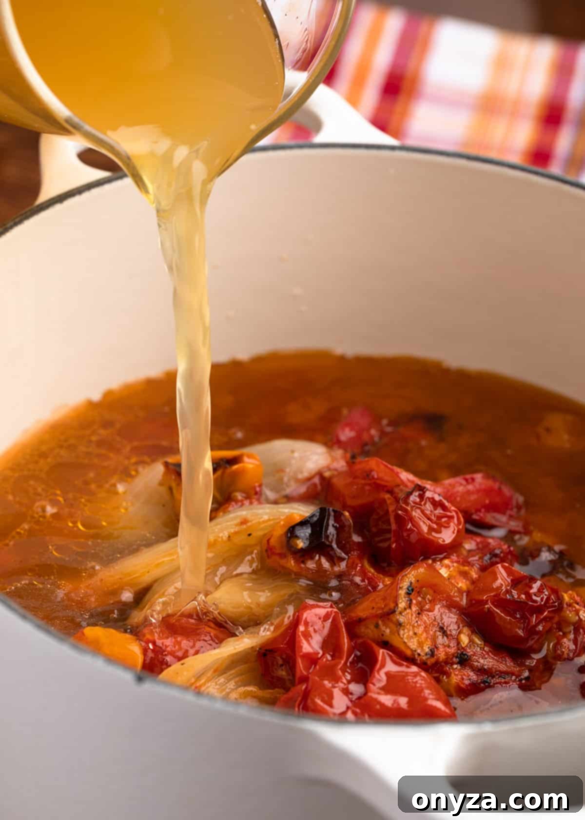 pouring chicken stock into a white pot filled with roasted tomatoes, onions, and garlic